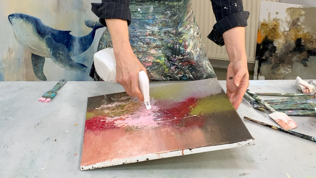 Eine Leinwand die mit Erde als Struktur grundiert wurde und jetzt beige, Braun, Grün und Rosa Acryl fliesst, eine Hand sprüht Wasser