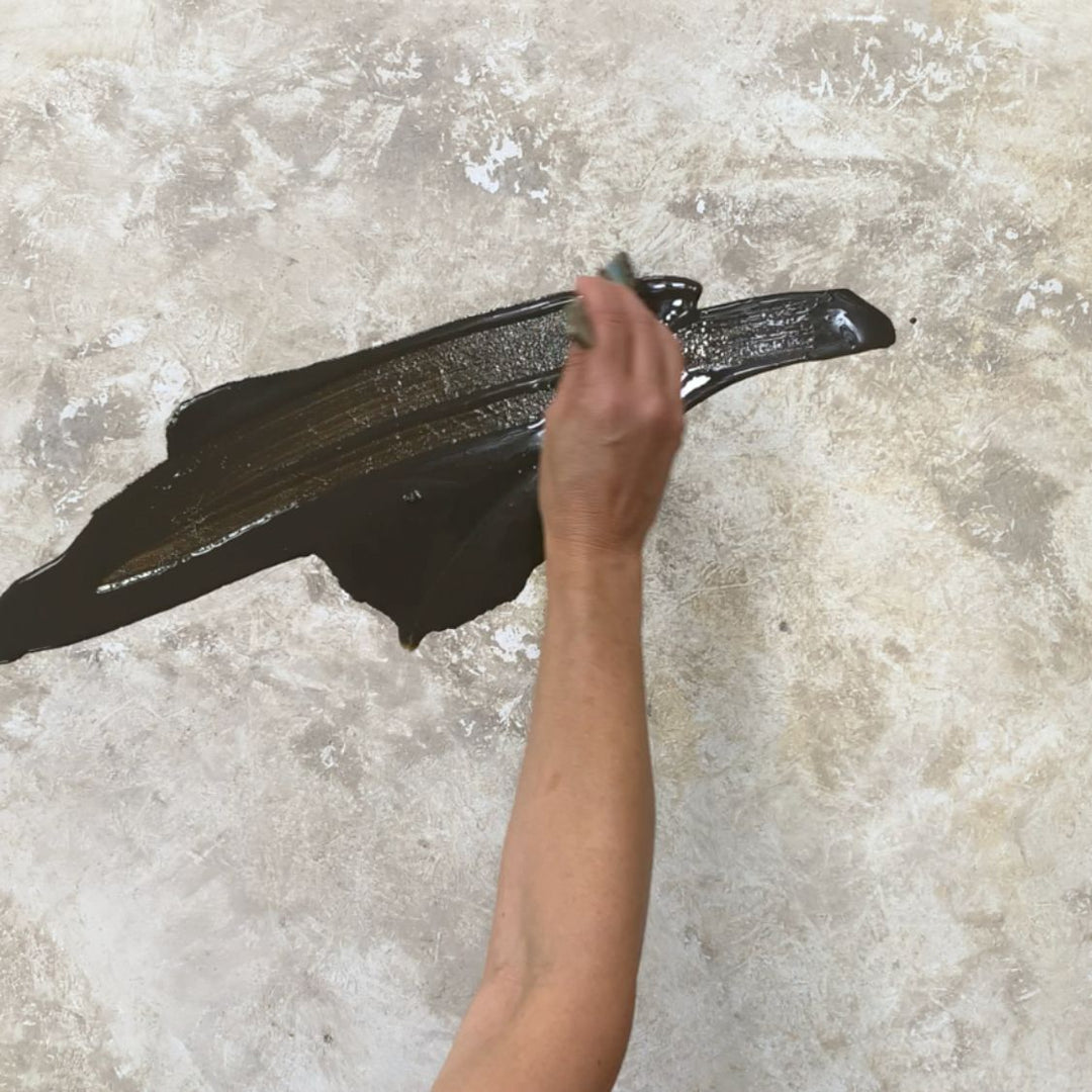 Hand zieht mit einer Spachtel einen breiten Strich in Dunkelbraun-Schwarz über eine strukturierte, helle Leinwand – Maltechnik Schichten und Struktur