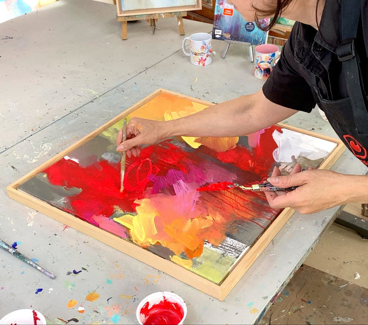 Petra Thölken malt mit Pinsel an einem abstrakten Acrylbild in Rot, Pink und Gelb auf einer gerahmten Holzplatte im Atelier.