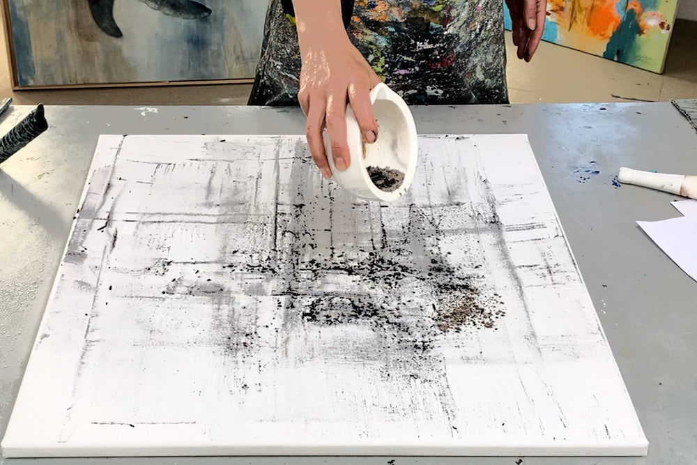 Petra Thölken streut dunkles Pigment auf eine weiße Leinwand mit schwarzer Vorzeichnung – Beginn eines abstrakten Malprozesses im Atelier.